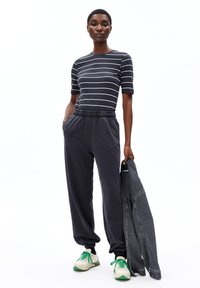 Vrouw in een zwart-wit gestreepte korte mouw top, donkere joggerbroek, witte sneakers met groene veters, en houdt een zwarte spijkerjack vast.