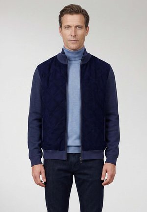 Man draagt een navy doorgestikte jas met ritssluiting over een lichtblauwe coltrui met donkere jeans, staand tegen een effen achtergrond.