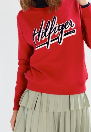 Czerwona bluza z logo "Hilfiger Denim" noszona na czarnym topie, w połączeniu z jasnozieloną plisowaną spódnicą.