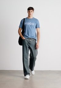 Μπλε βαμβακερό μπλουζάκι με λογότυπο "HUGO", συνδυασμένο με χαλαρές γκρι τζιν παντελόνες και λευκά αθλητικά παπούτσια. Το μοντέλο κρατάει μια μαύρη τσάντα. Απλός φόντος.