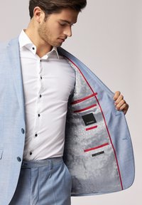 Hellblaues Sakko aus strukturiertem Stoff, mit einem gemusterten Futter, roten Akzenten an den Taschen und einem weißen Hemd mit schwarzen Knöpfen darunter.