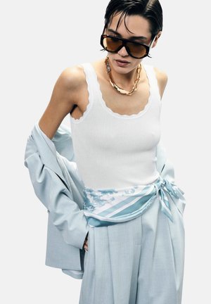 Persona che indossa occhiali da sole marroni oversize, una collana spessa a catena dorata, un top bianco smerlato, un blazer azzurro chiaro e pantaloni abbinati con cintura a sciarpa.