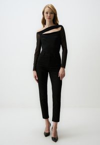 Schwarzer Bodysuit mit durchsichtigen langen Ärmeln, der einen asymmetrischen Ausschnitt hat. Kombiniert mit hochgeschnittenen schwarzen Jeans und schwarzen High Heels.