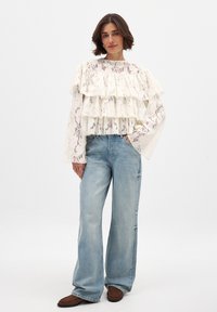 Cream lace top with layered ruffles, paired with high-waisted light blue denim jeans. Model stands on a plain background.