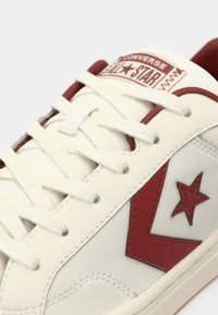White leather sneaker with red accents, including a star and logo, ample lacing, textured surface, and a low-profile design.