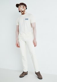 T-shirt crem cu logo albastru "Lee", asortat cu blugi albi ajustați. Șapcă maro și adidași bej deschis cu accente maro închis.
