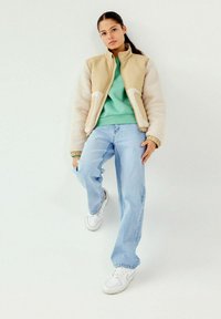Chaqueta cremosa y beige con cremallera y mangas de borreguito, combinada con un sudadera verde y vaqueros de azul claro. Zapatillas blancas complementan el conjunto.