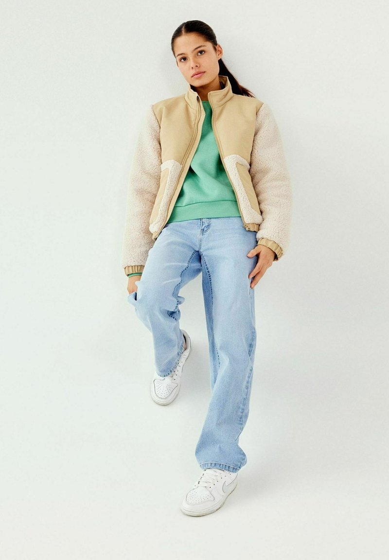 Chaqueta cremosa y beige con cremallera y mangas de borreguito, combinada con un sudadera verde y vaqueros de azul claro. Zapatillas blancas complementan el conjunto.