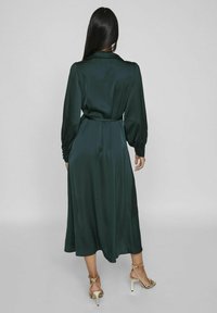 Abito in satin verde scuro con maniche lunghe, colletto e cintura da annodare. Caratterizzato da una silhouette fluida e dettagli a bottoni sui polsini.