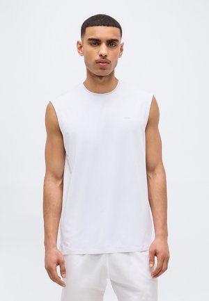 Νέος άνδρας με κοντά μαλλιά, φορώντας λευκό αμάνικο πουκάμισο και λευκό παντελόνι, στέκεται μπροστά σε απλό φωτεινό φόντο.