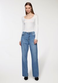 Biała bluzka z długimi rękawami połączona z niebieskimi jeansami o wysokim stanie i luźnym kroju. Zauważalne detale: dekolt w serek, tekstura denimu oraz ciemne buty na obcasie.