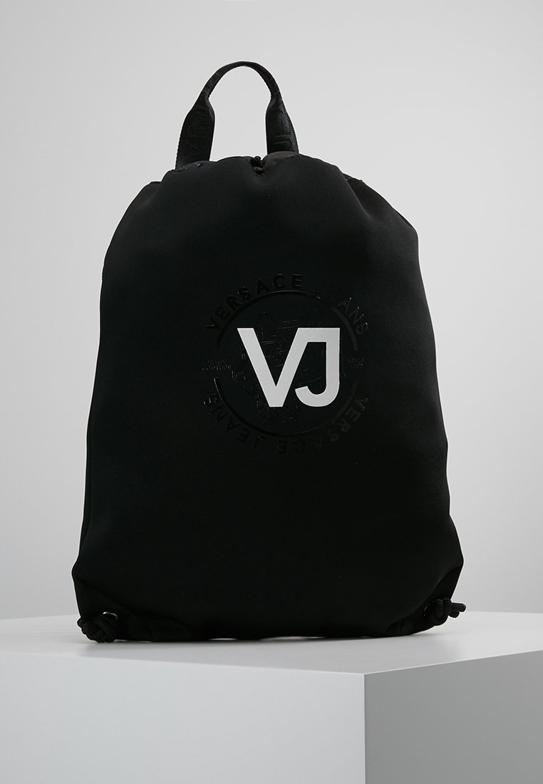 Sac à dos noir à cordon avec deux poignées, grand logo blanc "VJ" et marquage en relief "Versace Jeans" sur le devant, posé sur un piédestal blanc.