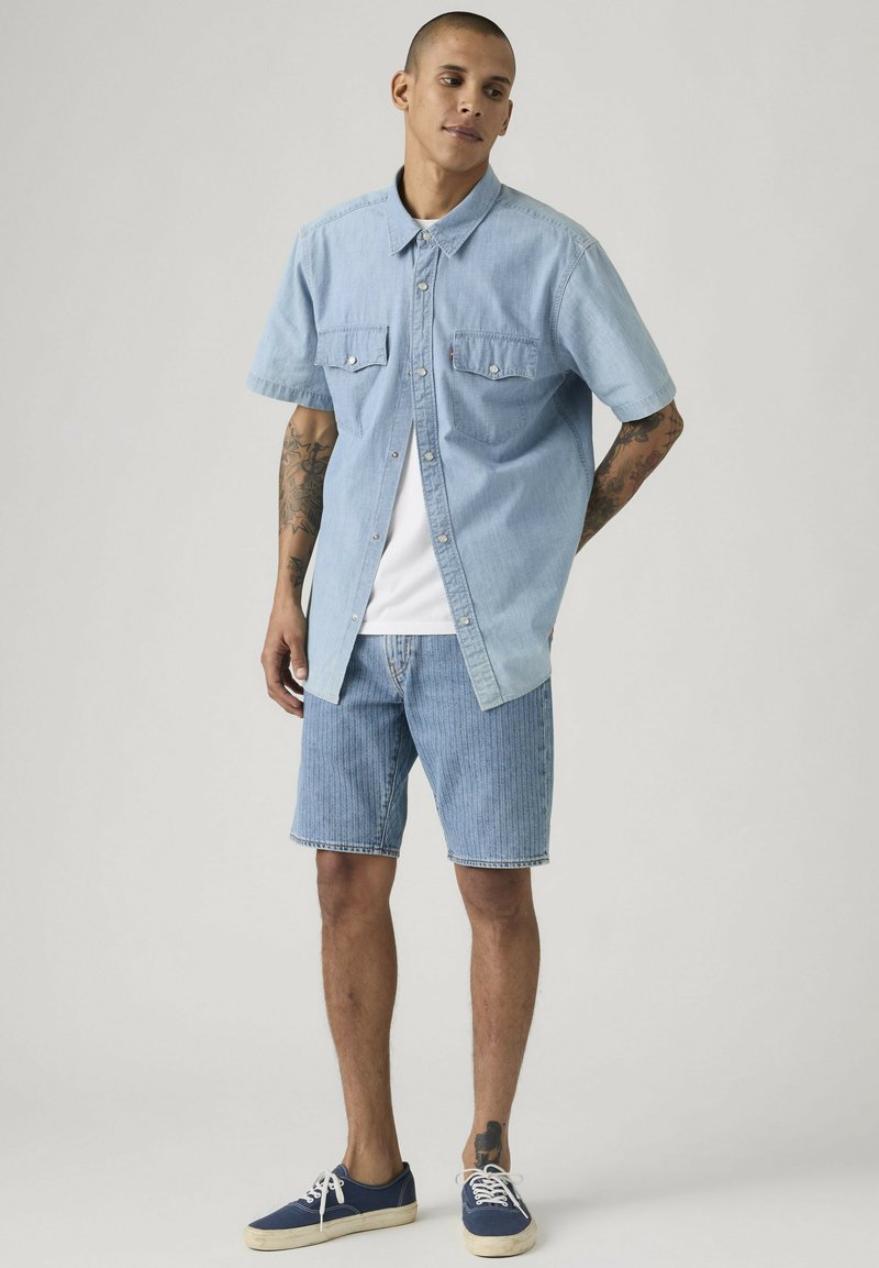 Hombre con cabello corto que lleva una camisa de mezclilla de manga corta azul claro, camiseta blanca, pantalones cortos de mezclilla y zapatillas deportivas azul marino, de pie con las manos detrás de la espalda.