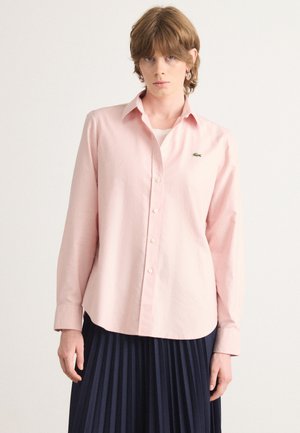 Chemise rose clair à boutons avec col pointu et manches longues, arborant un petit logo vert sur la poitrine, assortie à une jupe plissée bleu marine.