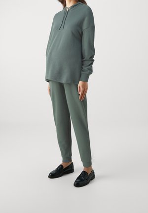 Kobieta w luźnej zielonej bluzie z kapturem i pasujących spodniach dresowych, ubrana w czarne mokasyny, stojąca na tle jednolitego jasnego tła.