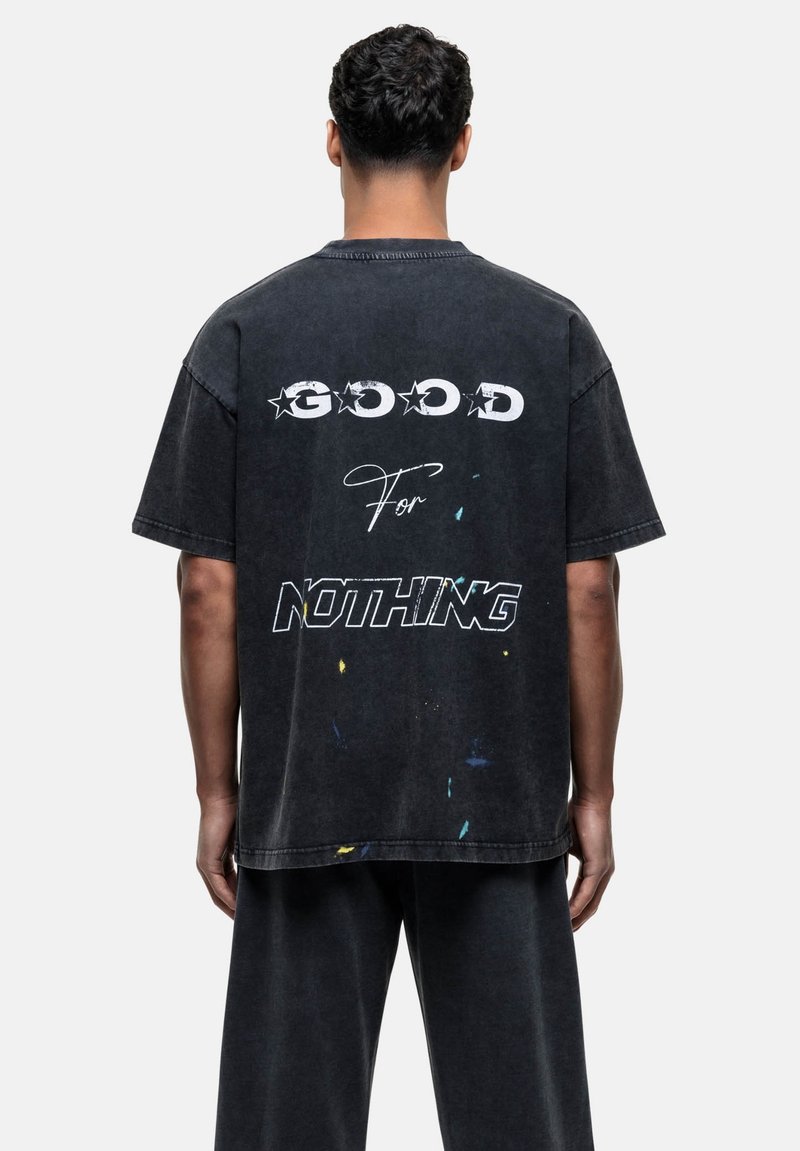 T-shirt noir oversize avec le texte graphique "GOOD For NOTHING" en blanc, et des éclaboussures de peinture colorées au dos. Design décontracté en coton.