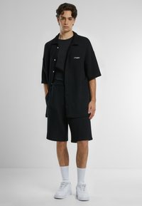 Svart, ruteret kortermet skjorte med knapper, parret med matchende shorts. Hvite sneakers og sokker fullfører antrekket. Minimalistisk design.
