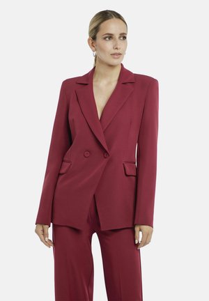 Woman wearing a fitted double-breasted burgundy blazer and matching pants, standing against a plain light background, looking to the side.