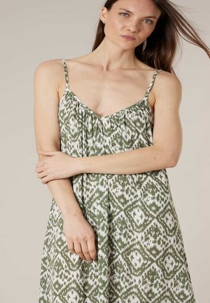 Frau mit langen braunen Haaren, die ein ärmelloses, grün-weiß gemustertes Kleid trägt und vor einem einfarbigen beigefarbenen Hintergrund steht.