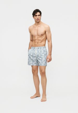 Man zonder shirt met kort donker haar, gekleed in blauw-wit patroon zwemshorts, staat op blote voeten met handen in de zakken tegen een effen achtergrond.