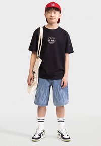 Svart t-skjorte med teksten "Utenom DENNE VERDEN", blå denimshorts med flammemønster, hvite sokker med svarte striper, og svarte joggesko.