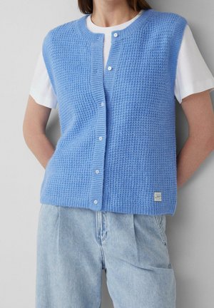 Kvinde iført en blå knappet strikvest over en hvid kortærmet skjorte og lyseblå jeans, stående med hænderne bag ryggen.