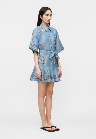 Γυναίκα που στέκεται με μπλε φόρεμα με φλοράλ σχέδιο, ζώνη στη μέση, κοντά μανίκια και μαύρα σαγιονάρα, μπροστά σε λευκό φόντο.