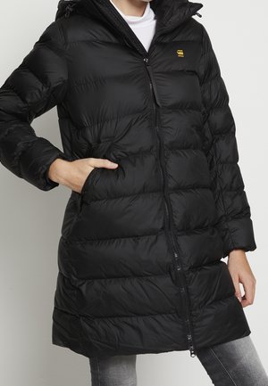 Femme portant un manteau rembourré noir avec fermeture éclair et capuche, une main dans la poche du manteau, associé à un jean gris.