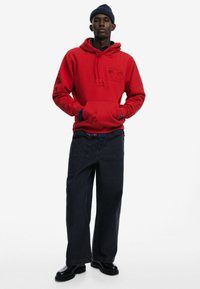 Červená mikina s kapucňou a predným vreckom, ozdobená grafickými dizajnmi, v kombinácii s tmavými širokými nohavicami a čiernymi čižmami, prezentovaná na neutrálnom pozadí.