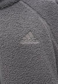 Lähedalt vaadates halli tekstuuriga fliiskangast, millel on brodeeritud Adidas'i logo, kolm triipu ja kaubamärgi nimi.
