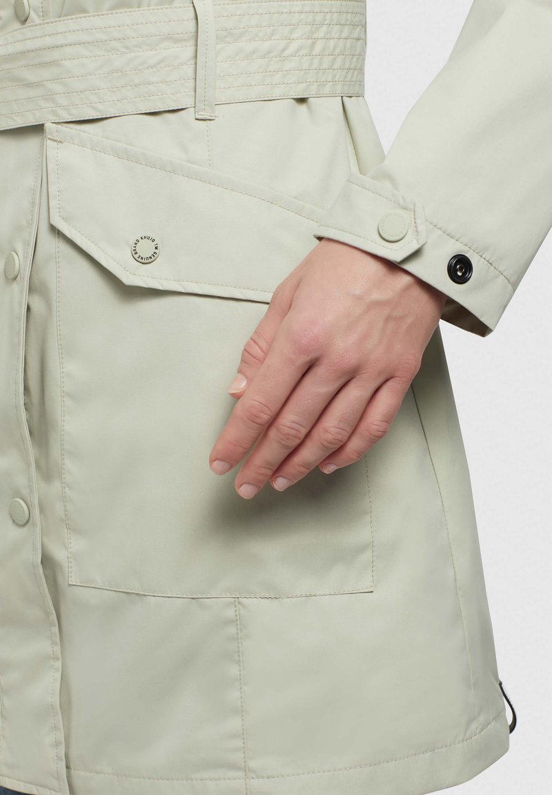 Giacca di colore chiaro, resistente all'acqua, con vita regolabile da cintura, grandi tasche e dettagli con bottoni; texture liscia e vestibilità su misura.