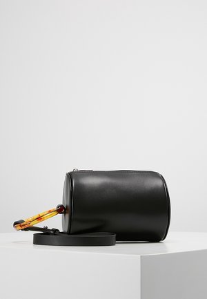 Bolso cilíndrico de cuero negro con un asa circular de carey y una correa de hombro negra desmontable sobre una superficie blanca.