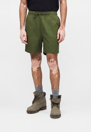 Άτομο που φοράει ελιά πράσινες σορτς, γκρι κάλτσες και μπότες σε χρώμα ώχρας με κορδόνια, στέκεται μπροστά σε ένα απλό φωτεινό φόντο.