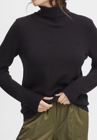 Svart polotröja med ett texturerat stickmönster, ribbad krage och manschetter, kombinerad med lätta olivgröna byxor.