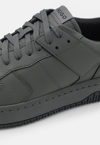 Gray leather sneaker featuring a textured surface, rounded toe, flat gray laces, and a black rubber sole. "HUGO" branding on the tongue.