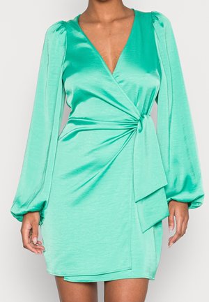 Frau trägt ein tealfarbenes Satin-Wickelkleid mit langen, puffigen Ärmeln und seitlicher Knotendetail, steht vor einem schlichten hellen Hintergrund.