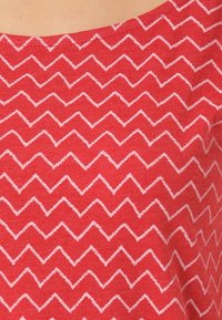 Czerwony materiał w białą zygzakowatą kratkę. Materiał wydaje się miękki o gładkiej fakturze. Dekolt jest szeroki i zaokrąglony.