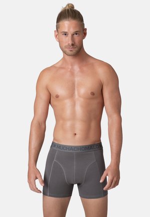 Grijze boxershorts met contrasterende zwarte tailleband waarop de merknaam "MUCHACHO MALO" staat, met een aangesloten ontwerp en een gladde textuur.