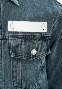 Chaqueta de mezclilla en azul con un lavado vintage texturizado. Presenta una etiqueta de cuero blanco con dos botones y un botón metálico con logo en el bolsillo del pecho.