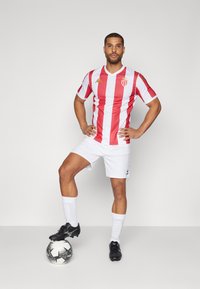 Rødt og hvitt stripete fotballtrøye med en sportslogo, paret med hvite shorts. Svarte fotballsko og en fotball ved føttene.