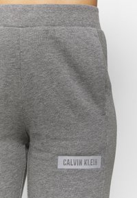 Pantalones de chándal de algodón grises con un acabado texturizado, que presentan un parche con el logo blanco que dice "CALVIN KLEIN" en el costado. Cintura elástica.