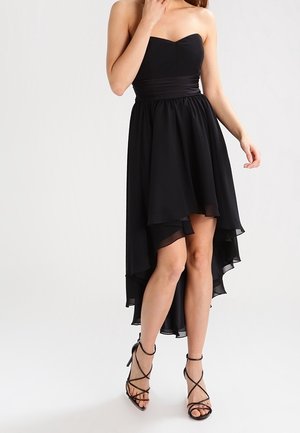 Robe noire bustier à taille froncée, modèle asymétrique (plus courte devant et plus longue derrière), portée avec des sandales noires à talons hauts et brides, sur une femme debout devant un fond blanc.