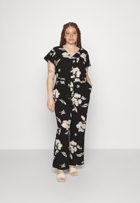 Svart blomstermønstrete jumpsuit med korte ermer, v-hals og belte i midjen. Har store hvite blomster og lommer. Brukt med lyse sandaler.