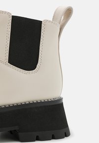 Gros plan sur une botte beige pour cheville avec panneau latéral élastique noir, semelle épaisse à crampons noire et languette de traction à l'arrière.