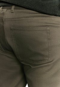 Ciemnooliwkowe spodnie o szczotkowanej fakturze, z jedną tylną kieszenią i subtelnymi detalami szycia. Prosty krój nogawki.