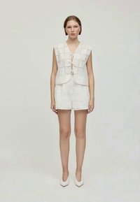 Chaleco blanco de eyelet con cierre frontal atado, combinado con pantalones cortos a juego. Textura suave, forma estructurada y patrón de recortes geométrico.