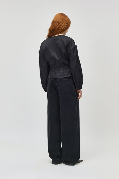Femme aux longs cheveux roux, de dos, portant un haut noir à manches longues et un jean noir à jambes larges sur un fond uni.
