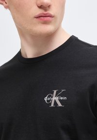 Νεαρός άνδρας που φοράει μαύρο μπλουζάκι με στρογγυλή λαιμόκοψη και μικρό ανοιχτό γκρι λογότυπο Calvin Klein στο αριστερό μέρος του στήθους.