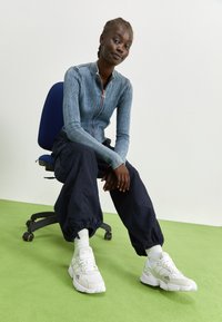 Tajt, ribbat blå zip-up skjorta, marina baggy byxor och vita sneakers med texturerade sulor. Sitter på en svart kontorsstol mot en grön golv.