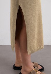 Abito lavorato a maglia beige con uno spacco laterale, caratterizzato da dettagli a costine sull'orlo. Indossato con sandali in scamosciato marrone con fibbia su una superficie grigia texturizzata.